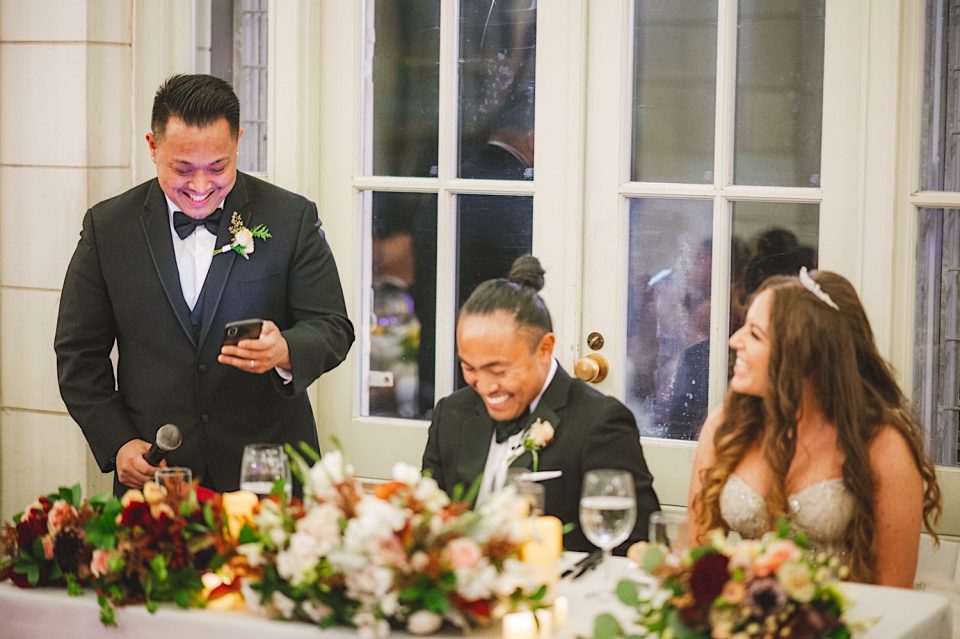 A man holding a microphone smiles while reading from his smartphone, sitting at an NYC wedding table with a laughing couple.