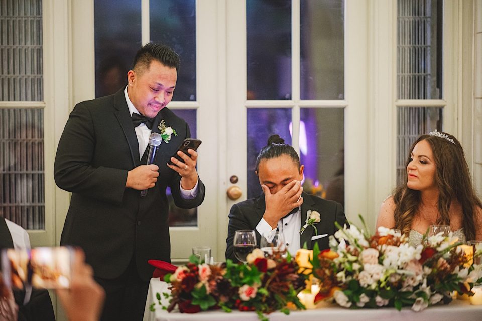 A man in a tuxedo reads from a smartphone at the Ash + John Boathouse NYC Wedding, standing beside another emotional man and a smiling woman in a tiara, with a flower-decorated table in the foreground.