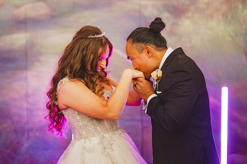 Ash and John kissing at their wedding reception in the Boathouse NYC, both dressed formally, under colorful lights with a soft purple haze in the background.