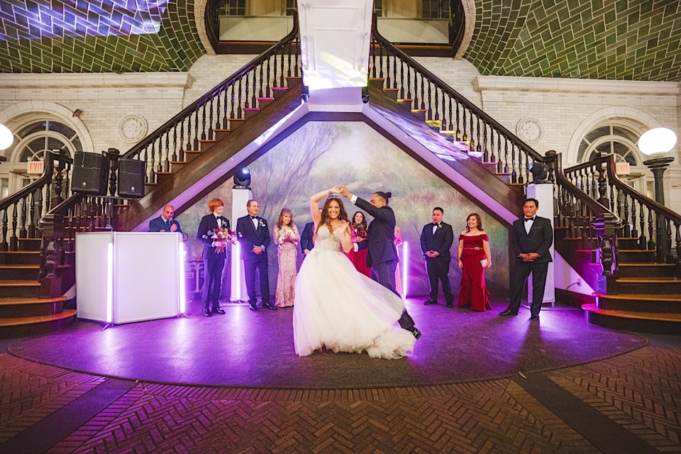 Bride dancing gracefully at her NYC wedding reception in a grand boathouse with a staircase, surrounded by guests and illuminated by soft purple lights.