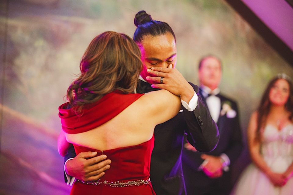 An emotional moment at a boathouse wedding reception with a woman in a red dress embracing a teary-eyed man in a suit, while others watch in the background.