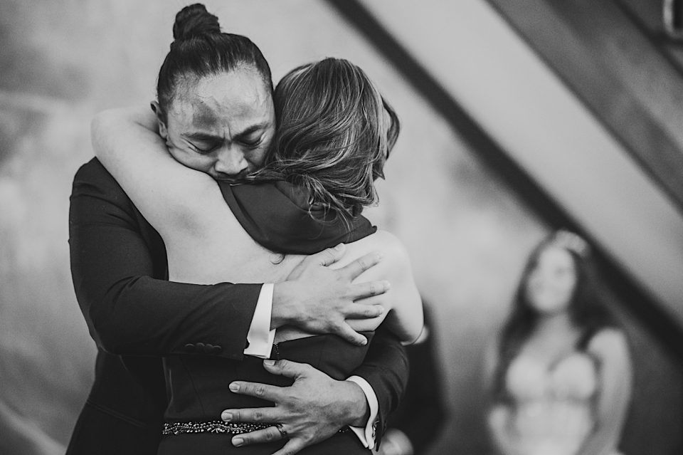 A man with a ponytail in a suit and a woman with short hair embrace tightly at their NYC wedding, expressing emotion, in black and white.