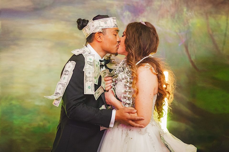A bride and groom, Ash and John, kiss passionately, adorned in money-themed accessories, against a soft, multicolored backdrop.