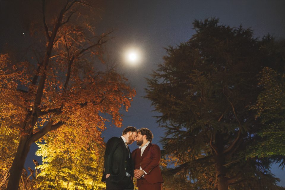 Two men in suits stand close together under a full moon, surrounded by tall autumn trees at night—captured as if ready for a Canon R1 review, with remarkable clarity and depth.