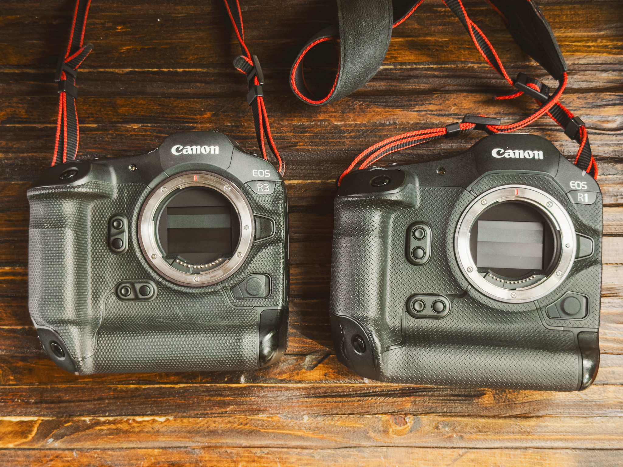 Two Canon DSLR cameras, models EOS R3 and EOS R1, sit side by side on a wooden surface with red and black straps attached—perfect for a detailed Canon R1 review.