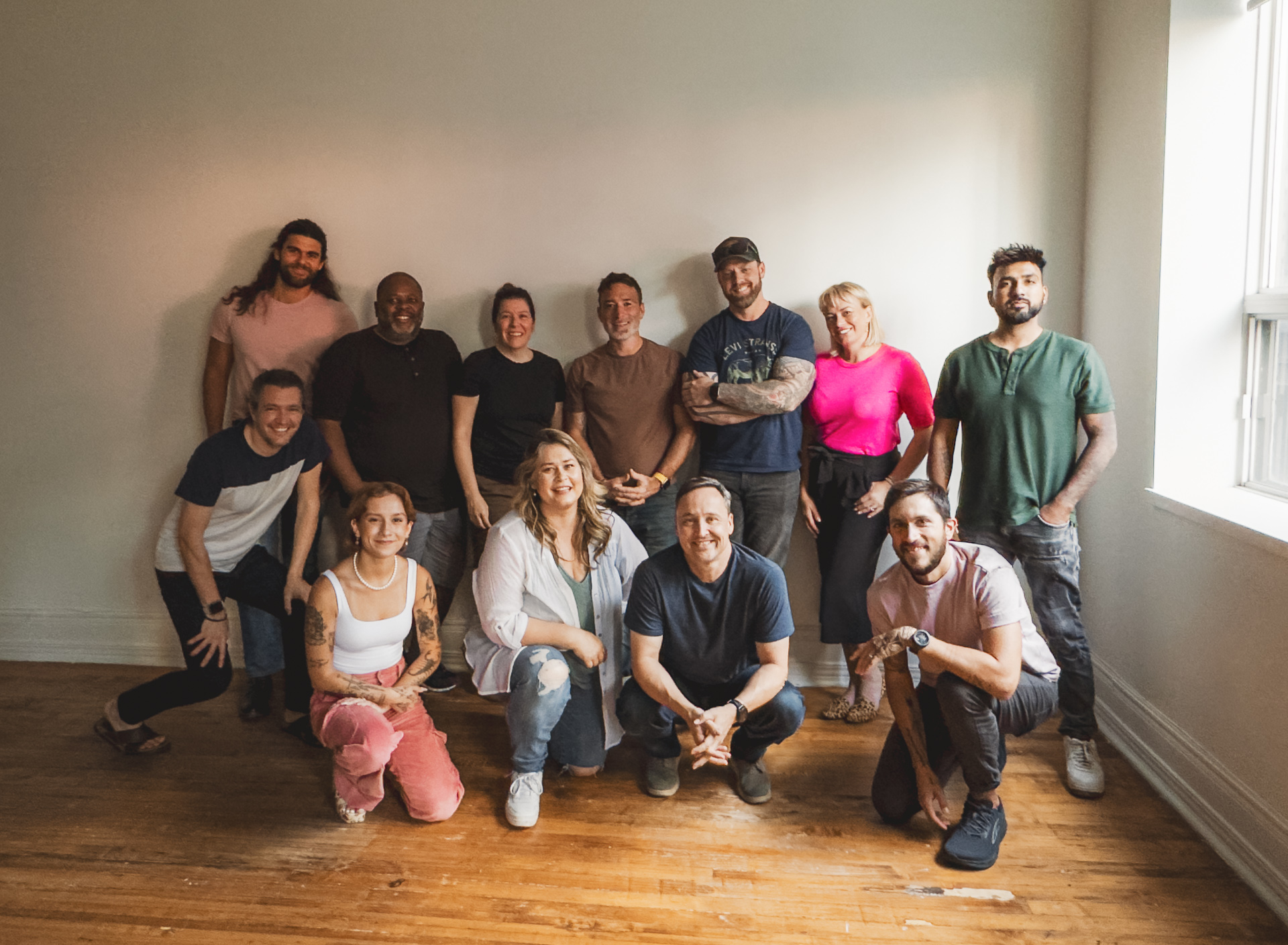 A group of 12 people of various ages stands and kneels in a well-lit room with a plain background, casually capturing an auto draft moment. Some are smiling, while others have neutral expressions. Everyone is dressed in casual clothing.