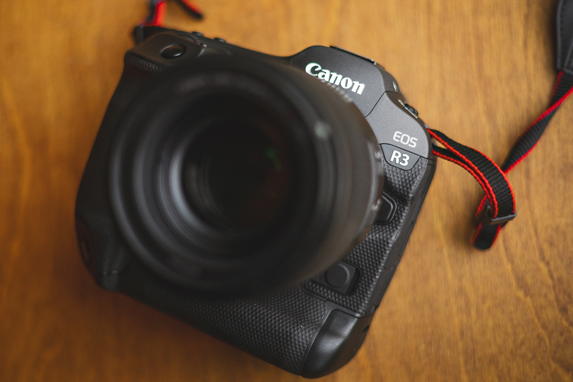 Close-up of a Canon EOS R3 camera with a lens attached, placed on a wooden surface. The camera strap with a red edge is partially visible, ready for an auto draft photography session.